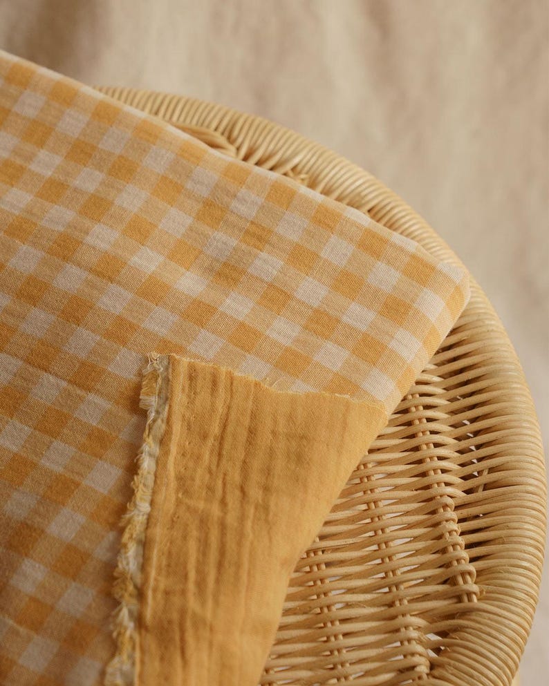 May include: Close-up of a folded fabric with a yellow and white gingham pattern. The fabric is resting on a woven wicker surface. The reverse side of the fabric is a solid yellow color, with a frayed edge.