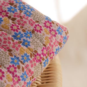 May include: A quilted fabric item featuring a repeating floral pattern on a light pink background. The design includes small blue, magenta, and yellow flowers interspersed with brown leaves and textured beige circular motifs across the surface.