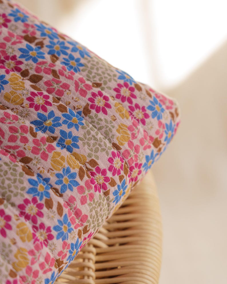 May include: A quilted fabric item featuring a repeating floral pattern on a light pink background. The design includes small blue, magenta, and yellow flowers interspersed with brown leaves and textured beige circular motifs across the surface.