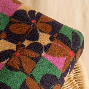 May include: Close-up of a patterned fabric with a floral design. The fabric features large, stylized flowers in brown, navy, and tan, set against a backdrop of green and pink squares. The flowers have red centers.