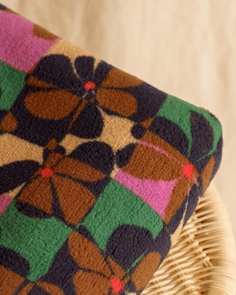 May include: Close-up of a patterned fabric with a floral design. The fabric features large, stylized flowers in brown, navy, and tan, set against a backdrop of green and pink squares. The flowers have red centers.