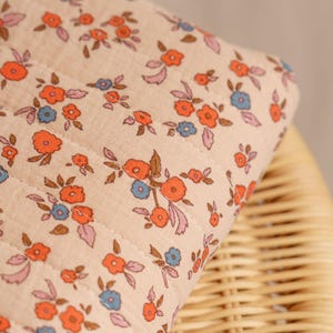 May include: Close-up of a quilted fabric with a floral pattern. The fabric features small orange, blue, and pink flowers with brown leaves on a light beige background. The fabric is quilted with vertical stitching.
