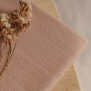 May include: A folded, dusty rose-colored fabric with a geometric pattern. The fabric is draped over a light-colored surface, and dried flowers are placed on the left side of the fabric.
