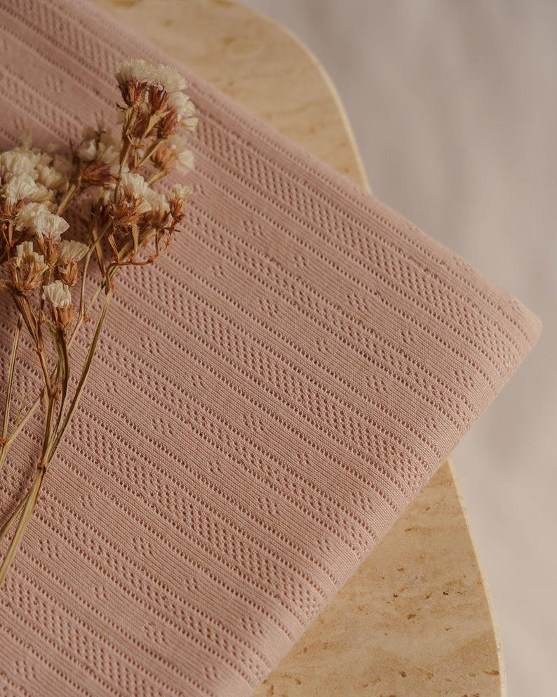 May include: A folded, dusty rose-colored fabric with a geometric pattern. The fabric is draped over a light-colored surface, and dried flowers are placed on the left side of the fabric.