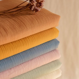 Pode incluir: Uma pilha de tecido dobrado em várias cores pastel, incluindo pêssego, amarelo, azul, rosa, verde e branco. Um raminho de flores secas repousa no topo. O tecido parece ser um material leve e texturizado.