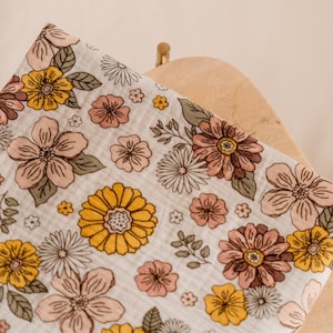 May include: A white muslin swaddle blanket with a floral print in shades of yellow, pink, and brown. The flowers are stylized and have a retro feel.