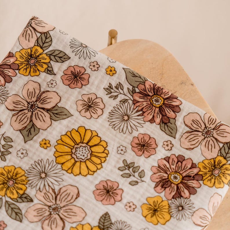 May include: A white muslin swaddle blanket with a floral print in shades of yellow, pink, and brown. The flowers are stylized and have a retro feel.