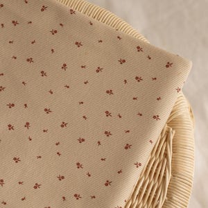 Könnte beinhalten: Ein gefaltetes Stück beigen Stoffes mit einem sich wiederholenden Muster aus kleinen, dunkelroten floralen Designs. Der Stoff liegt auf einem geflochtenen Korbstuhl mit neutralem Hintergrund.