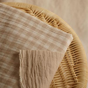 May include: Close-up of two pieces of fabric in shades of beige and tan. One piece has a checkered pattern, while the other has a textured, wrinkled appearance. The fabrics are draped over a woven wicker chair.
