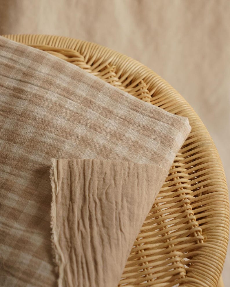 May include: Close-up of two pieces of fabric in shades of beige and tan. One piece has a checkered pattern, while the other has a textured, wrinkled appearance. The fabrics are draped over a woven wicker chair.