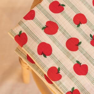 Peut inclure: Un textile plié présentant un motif répétitif de pommes rouge vif avec de petites tiges vertes. Le tissu possède un fond crème clair accentué par de fines rayures verticales vertes courant parallèlement aux motifs de pommes sur la surface.