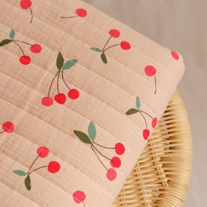 May include: A light pink, quilted fabric with a repeating pattern of red cherries and green leaves. The fabric is arranged on a woven wicker chair, showcasing the texture and design of the textile. The cherries are in various stages of ripeness.