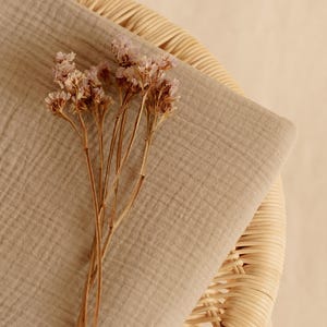 Op de afbeelding: Een gevouwen, lichtbruine stof met een getextureerd oppervlak, mogelijk linnen of katoen, rust op een geweven rieten stoel. Delicate gedroogde bloemen met lichtpaarse bloesems en bruine stelen zijn op de stof geplaatst, waardoor een natuurlijke, neutraal getinte compositie ontstaat.