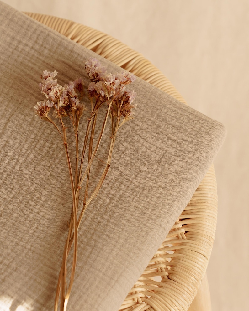 Op de afbeelding: Een gevouwen, lichtbruine stof met een getextureerd oppervlak, mogelijk linnen of katoen, rust op een geweven rieten stoel. Delicate gedroogde bloemen met lichtpaarse bloesems en bruine stelen zijn op de stof geplaatst, waardoor een natuurlijke, neutraal getinte compositie ontstaat.