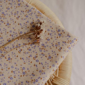 May include: A folded piece of fabric with a cream-colored ribbed texture, adorned with a delicate floral pattern in shades of purple and brown. A dried flower stem rests on top. The fabric is likely used for sewing or crafting.
