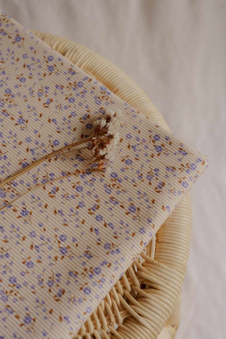 May include: A folded piece of fabric with a cream-colored ribbed texture, adorned with a delicate floral pattern in shades of purple and brown. A dried flower stem rests on top. The fabric is likely used for sewing or crafting.