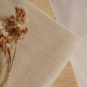 May include: A folded, cream-colored fabric with a textured, patterned design. The fabric is draped over a light-colored, geometric surface. Dried flowers are placed on the left side of the fabric.