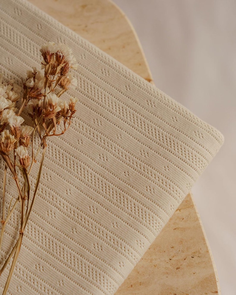 May include: A folded, cream-colored fabric with a textured, patterned design. The fabric is draped over a light-colored, geometric surface. Dried flowers are placed on the left side of the fabric.