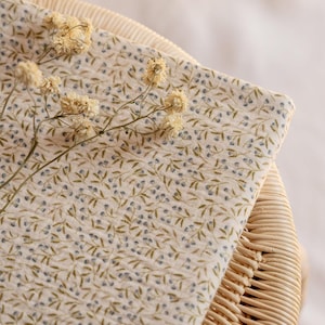 以下が含まれることがあります： クリーム色の背景に、青いベリーと緑の葉の繊細な花柄が施された折り畳まれた布。織り込まれた籐の表面に布が置かれ、その上にドライフラワーが置かれています。