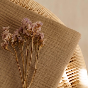 Op de afbeelding: Een close-up toont een beige stof die over een rieten stoel is gedrapeerd. Gedroogde lavendelkleurige bloemen liggen op de stof. De sc&egrave;ne wordt verlicht door zacht, natuurlijk licht, wat een warme uitstraling cre&euml;ert. De stof lijkt linnen of een vergelijkbaar natuurlijk materiaal te zijn.