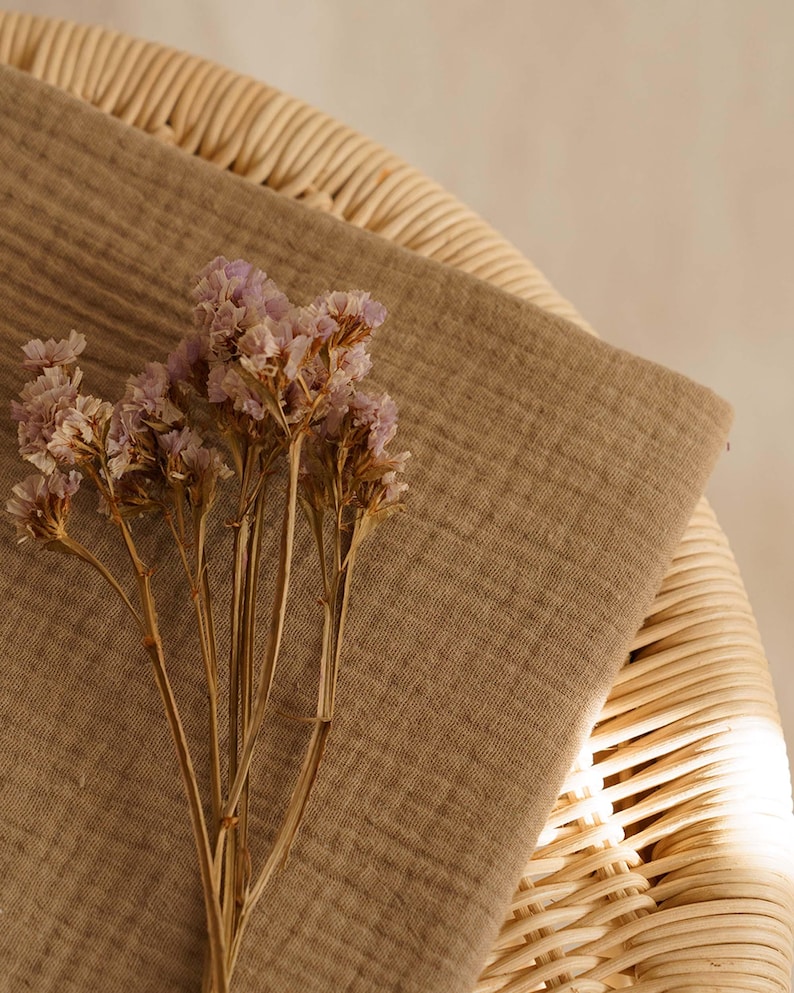 Op de afbeelding: Een close-up toont een beige stof die over een rieten stoel is gedrapeerd. Gedroogde lavendelkleurige bloemen liggen op de stof. De sc&egrave;ne wordt verlicht door zacht, natuurlijk licht, wat een warme uitstraling cre&euml;ert. De stof lijkt linnen of een vergelijkbaar natuurlijk materiaal te zijn.