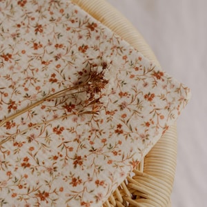 May include: A folded piece of fabric with a floral pattern in shades of brown and pink, resting on a woven wicker chair. The fabric has a cream-colored background with small, detailed flowers and leaves. A dried flower stem lies across the fabric.