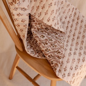 May include: A quilted textile featuring a cream-colored base with a repeating pattern of small brown floral sprigs. The fabric displays a vertical striped texture and a folded corner revealing a secondary brown and cream floral print pattern.
