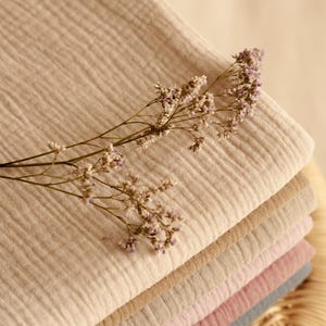 Op de afbeelding: Een stapel gevouwen stof in verschillende neutrale kleuren, waaronder beige, bruin en roze. Een takje gedroogde bloemen met kleine paarse bloemen ligt erop. De stof lijkt een zacht, lichtgewicht materiaal te zijn.