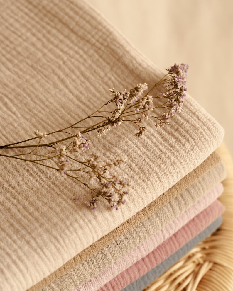Op de afbeelding: Een stapel gevouwen stof in verschillende neutrale kleuren, waaronder beige, bruin en roze. Een takje gedroogde bloemen met kleine paarse bloemen ligt erop. De stof lijkt een zacht, lichtgewicht materiaal te zijn.