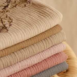 Op de afbeelding: Een stapel gevouwen, getextureerde stof in verschillende neutrale kleuren, waaronder beige, bruin, roze en grijs. De stof heeft een gekreukeld uiterlijk. Een takje gedroogde bloemen met paarse knoppen ligt bovenop de stof.