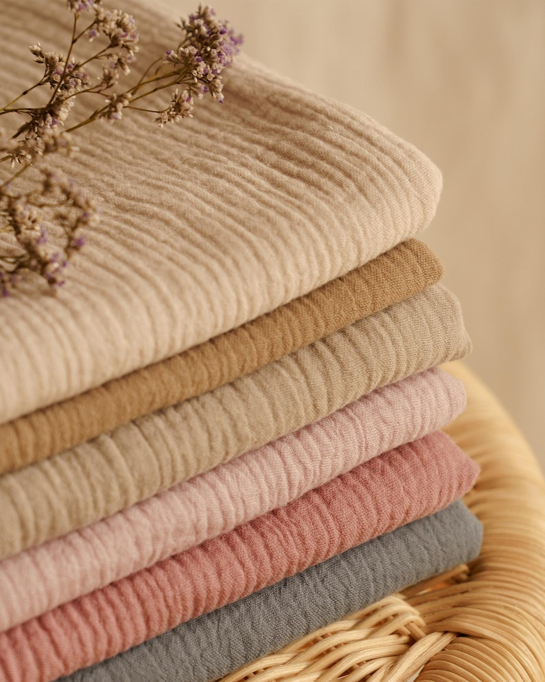 Op de afbeelding: Een stapel gevouwen, getextureerde stof in verschillende neutrale kleuren, waaronder beige, bruin, roze en grijs. De stof heeft een gekreukeld uiterlijk. Een takje gedroogde bloemen met paarse knoppen ligt bovenop de stof.