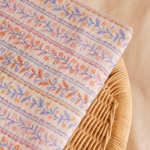 May include: A folded piece of fabric with a floral pattern in shades of peach, lavender, and blue on an off-white background. The fabric is resting on a woven wicker surface. The floral design includes delicate vines and small flowers.