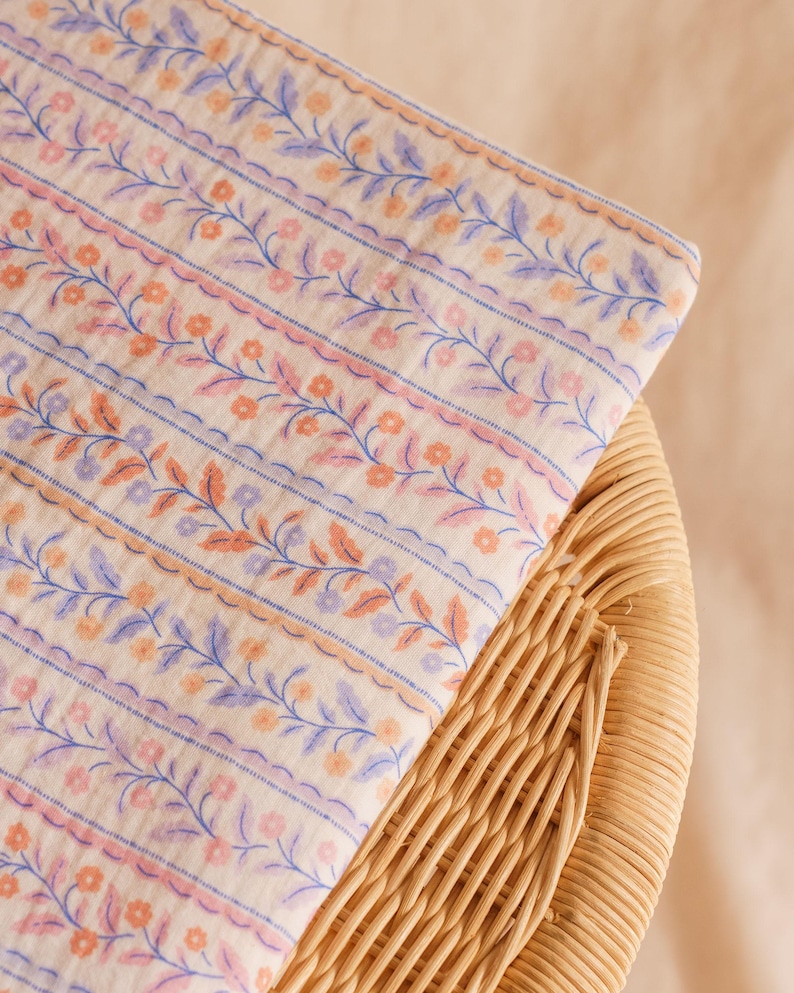 May include: A folded piece of fabric with a floral pattern in shades of peach, lavender, and blue on an off-white background. The fabric is resting on a woven wicker surface. The floral design includes delicate vines and small flowers.