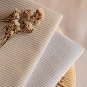 Può includere: Due pezzi di tessuto piegati con un motivo a righe sottili, uno in una tonalità beige tenue e l'altro in una tonalità azzurro chiaro. Un piccolo mazzo di fiori secchi color bianco sporco poggia sul tessuto beige, aggiungendo una consistenza naturale.