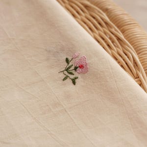 K&ouml;nnte beinhalten: Ein hellbeiger Stoff mit einer kleinen, feinen Stickerei einer rosa Blume mit gr&uuml;nen Bl&auml;ttern und St&auml;ngeln. Das Textil weist ein dezentes, strukturiertes Gewebe und ein weiches, mattes Erscheinungsbild auf, das die feine Handarbeit zeigt.