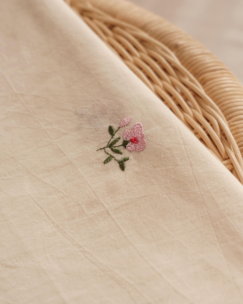 K&ouml;nnte beinhalten: Ein hellbeiger Stoff mit einer kleinen, feinen Stickerei einer rosa Blume mit gr&uuml;nen Bl&auml;ttern und St&auml;ngeln. Das Textil weist ein dezentes, strukturiertes Gewebe und ein weiches, mattes Erscheinungsbild auf, das die feine Handarbeit zeigt.