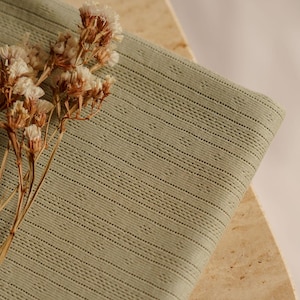 May include: Sage green fabric with a textured, striped pattern. The fabric is folded, revealing its detailed weave. Dried, light-colored flowers are placed on the fabric, adding a natural element to the composition. The fabric appears to be cotton or linen.