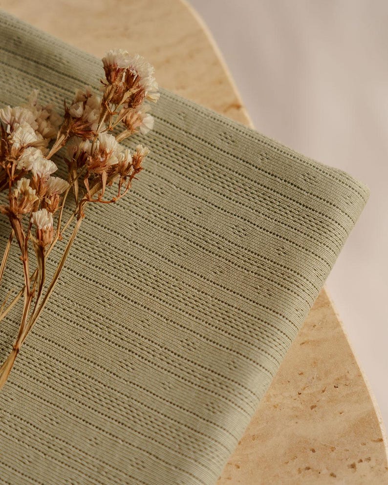 May include: Sage green fabric with a textured, striped pattern. The fabric is folded, revealing its detailed weave. Dried, light-colored flowers are placed on the fabric, adding a natural element to the composition. The fabric appears to be cotton or linen.