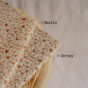 May include: Two fabric swatches, Muslin and Jersey, with a cream background and a delicate floral pattern in shades of brown and pink. The fabrics are folded over a woven wicker chair.