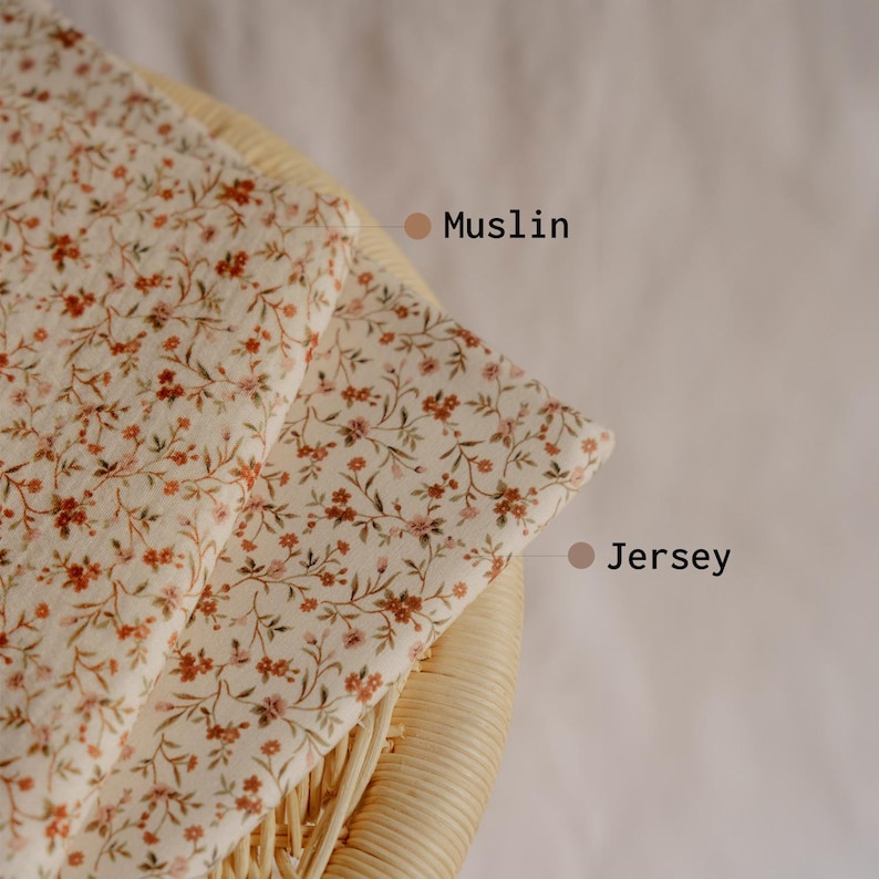 May include: Two fabric swatches, Muslin and Jersey, with a cream background and a delicate floral pattern in shades of brown and pink. The fabrics are folded over a woven wicker chair.