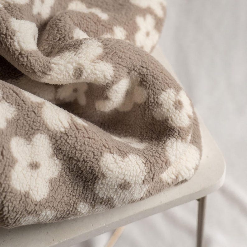 Puede incluir: Una manta suave y mullida con un patr&oacute;n floral blanco sobre un fondo beige.