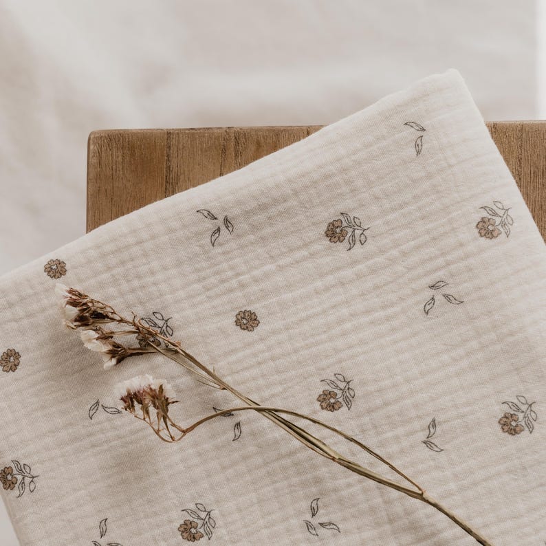 May include: Cream-colored muslin fabric with a floral pattern in brown and gray. The fabric is folded on a wooden surface, with a dried flower stem resting on top. The floral design includes small flowers and leaves.