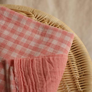 May include: A close-up shot of a pink and white gingham fabric, folded over a woven wicker basket. The fabric has a soft, textured appearance. The bottom edge of the fabric is a solid pink color. The basket is a light brown color.