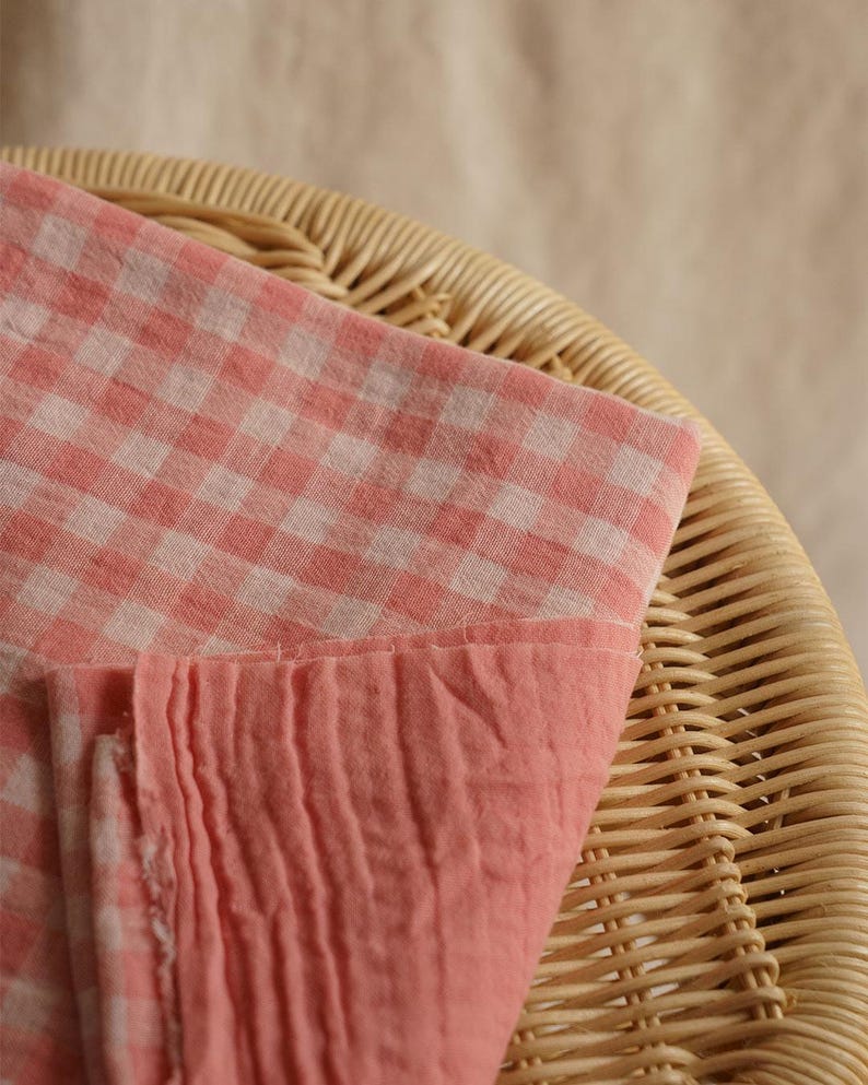 May include: A close-up shot of a pink and white gingham fabric, folded over a woven wicker basket. The fabric has a soft, textured appearance. The bottom edge of the fabric is a solid pink color. The basket is a light brown color.