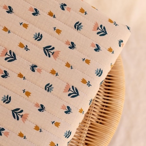 Può includere: Una coperta in tessuto trapuntato con un motivo ripetitivo di piccoli fiori simili a tulipani in tonalità rosa tenue, giallo senape e verde acqua scuro su uno sfondo beige chiaro. Il tessuto presenta linee verticali cucite sulla superficie.
