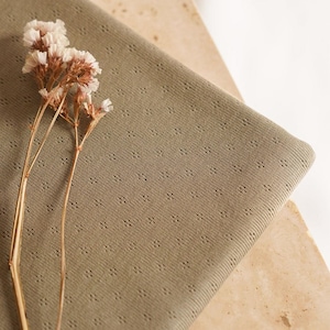 Op de afbeelding: Een close-up van een saliegroene stof met een subtiel textuurpatroon. De stof is gevouwen en gedrapeerd over een lichtbruin oppervlak. Een takje gedroogde witte bloemen is op de stof gelegd.