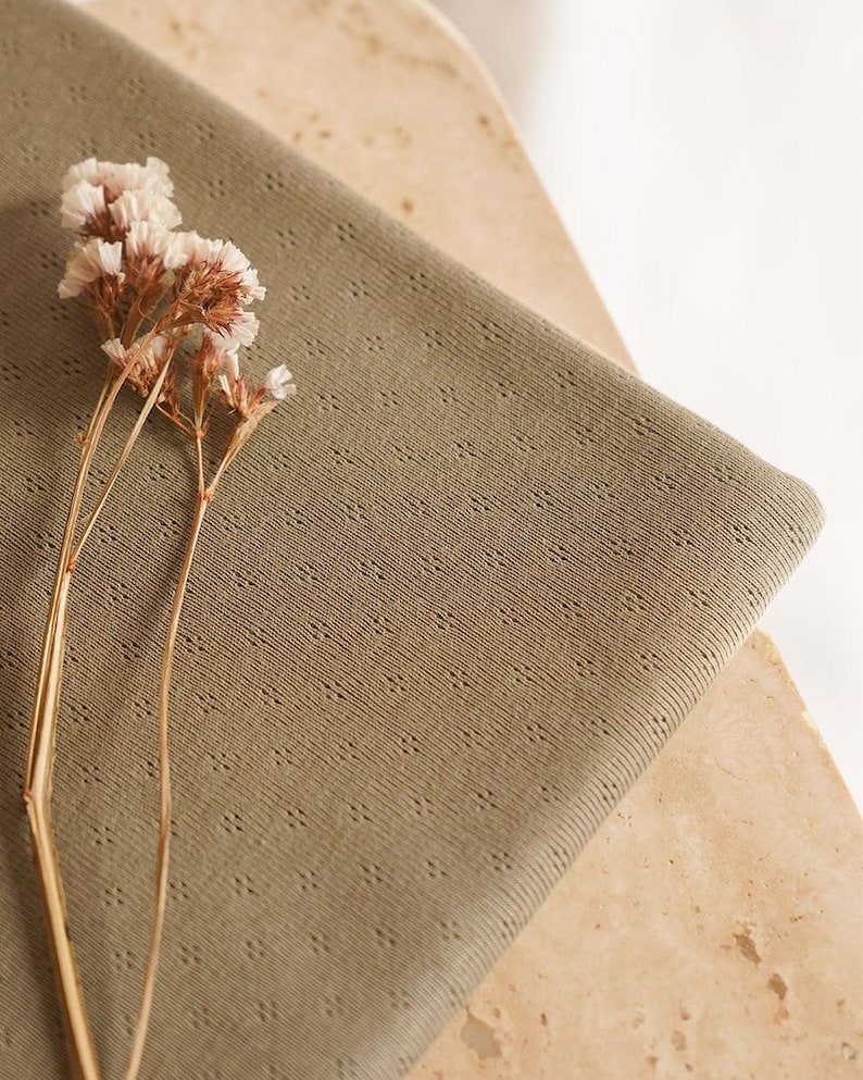 Op de afbeelding: Een close-up van een saliegroene stof met een subtiel textuurpatroon. De stof is gevouwen en gedrapeerd over een lichtbruin oppervlak. Een takje gedroogde witte bloemen is op de stof gelegd.