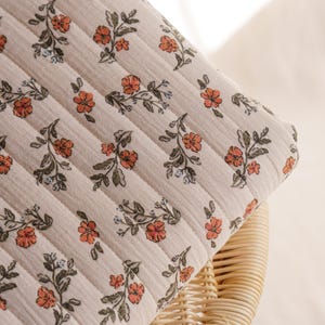 May include: Close-up of a cream-colored quilted fabric with a floral pattern. The pattern features orange flowers and green leaves with small blue accents. The fabric is folded over a wicker chair.