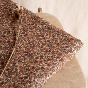 May include: A close-up of a beige fabric with a small-scale floral print in shades of brown and orange. The fabric is folded and draped over a light brown surface.