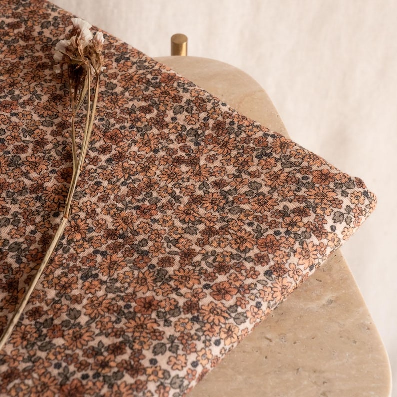 May include: A close-up of a beige fabric with a small-scale floral print in shades of brown and orange. The fabric is folded and draped over a light brown surface.